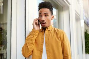 Portrait of young shocked dark skinned man in yellow shirt, walking down the street and speaks on the phone with his friends, hears unbelievable news, wit wide open mouth and eyes. photo