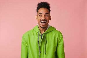 Close up of young happy amazed african american handsome guy broadly smiles, looks at the camera in surprise with wide open mouth, stands over pink background. photo