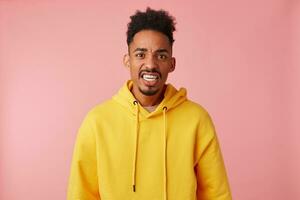 Portrait of young querulous african american guy in yellow hoodie, frowningin and misunderstanding of what is happening,ugly looking at the camera over pink background. photo