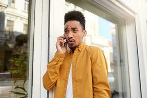 Young dissatisfied dark skinned man in yellow shirt, speaks on the phone with his friends and walking down the street, with resentful expression. photo