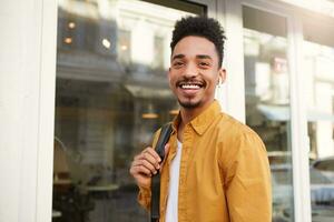 Close up of young broadly smiling dark skinned guy in yellow shirt walking down the street and looks cheerful, enjoy the sunny day in the city, listening favorite song on headphones. photo