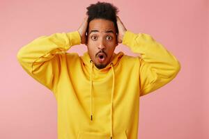 Young shocked african american guy in yellow hoodie, holding his head, his favorite football team missed a goal, looking at the camera with wide-open eyes and mouth, stands over pink background. photo