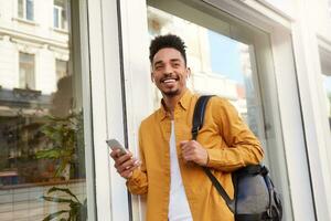 Young smiling African American guy in yellow shirt, looks happy and broadly smiling, walking down the street and holds telephone, enjoys his favorite song on headphones.. photo