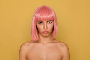 Offended young pretty blue-eyed lady with short pink bob haircut being upset about something and pounting her lips with sad face, standing over mustard background photo