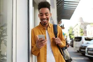 Young cheerful African American guy in yellow shirt, walking down the street and holds telephone, got a message with a funny video, looks joyful and broadly smiling. photo