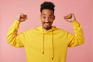 joven alegre africano americano hombre en amarillo sudadera, levantamiento su puños arriba, decir soy Frío' y soportes terminado rosado antecedentes y sonriente en general. foto