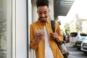 Young happy dark skinned man in yellow shirt, walking down the street and holds telephone, got a message with a funny video, looks joyful and broadly smiling. photo