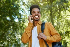 Young cheerful African American boy in yellow shirt, walking at the park, speaking on smartphone with his girlfriend, looking away and broadly smiling, enjoy the day. photo