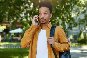 Portrait of young calm dark skinned man in yellow shirt walking on the park , holds telephone, waiting answer from his girlfriend, looks thoughtfully. photo