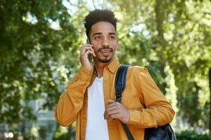 Young positive dark skinned man in yellow shirt, walking at the park and speaking on telephone, waitings his friends and looks away. photo