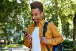 Young dissatisfied African American man in yellow shirt, chatting on the phone with his friends and walking at the park, looks at gadget with resentful expression, his friend late. photo