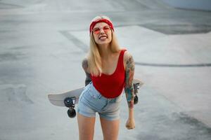 Nice blonde lady with tattooed arms dressed in a red T-shirt and denim shorts, with a knitted bandana on her head, in red glasses, holding a longboard in her hand and grinning. photo