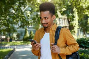 Young dissatisfied dark skinned guy, chatting on the phone with his friends and walking at the park, looking from under his forehead with resentful expression, his girlfriend is late again. photo