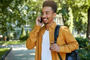 Young smiling dark skinned student walking at the park, speaking on smartphone, waitng his friend, wears in yellow shirt, looking away and broadly smiling. photo