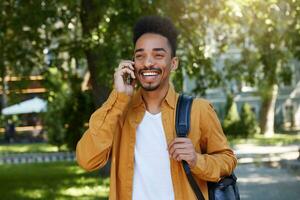 Young African American smiling boy walking afterr study in the park, talking on the phone, wears in a yellow shirt and a white T-shirt with a backpack on one shoulder, smiling and enjoys the day. photo