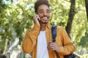 Photo of young African American glad man, walking at the park and talking on the phone with his friend, laugting and enjoying the day.