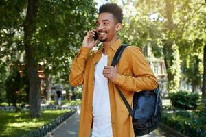 Young dark skinned smiling guy wears in a white shirt and a white T-shirt with a backpack on one shoulder, walking in the park and talking on the phone with his friend, smiling and enjoys the day. photo