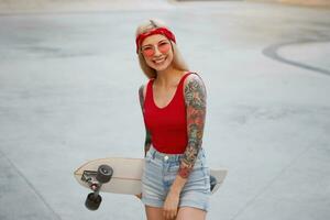 Cute blonde lady with tattooed arms in red glasses, red T-shirt and denim shorts with a knitted bandana on head, holding a longboard in hand, broadly smiling, spends time in skate park and enjoys. photo