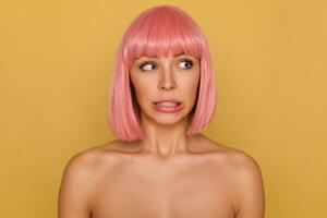 Confused young pretty blue-eyed pink haired female with bob haircut showing her teeth while twisning embarrassedly her mouth, isolated over mustard background photo