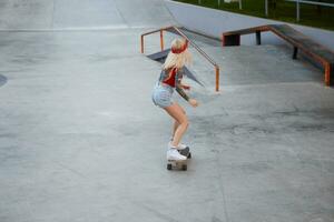 joven dama con rubia cabello, con tatuado brazos, usa en un rojo camiseta y mezclilla bermudas, con un de punto pañuelo en su cabeza, disfrutando longboard en patinar parque. foto