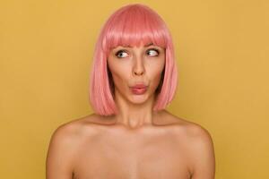 Close-up of excited young beautiful lady with short pink hair folding her lips while looking confusedly aside, posing over mustard background with naked shoulders photo