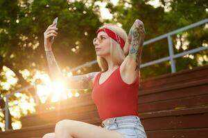 retrato de joven hermosa rubia mujer con tatuado brazos participación teléfono inteligente y hace selfie para su Blog. usa en un rojo camiseta y mezclilla bermudas, con un de punto pañuelo en cabeza, en rojo lentes. foto