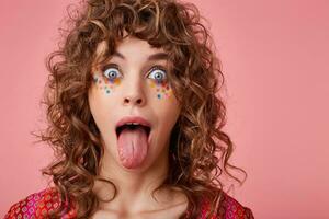 Poratrait of young curly blue-eyed woman with multicolored dots on her face looking at camera with wide eyes opened and sticking out her tongue, isolated over pink background photo