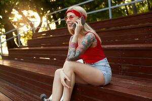 Young nice laughing blonde woman with tattooed arms, talking to his friend on smartphone, wears in red T-shirt, denim shorts, with knitted bandana on her head, in red glasses. photo