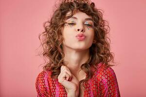 Cheerful young curly brunette female with festive hairstyle and multicolored dots on her face standing over pink background, folding lips in air kiss and looking joyfully to camera photo