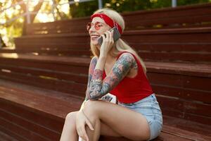 retrato de joven bonito rubia mujer con tatuado brazos, participación un teléfono inteligente a el oído y hablando a su amigo. orejas en rojo camiseta, mezclilla bermudas, con de punto pañuelo en su cabeza, en rojo lentes. foto