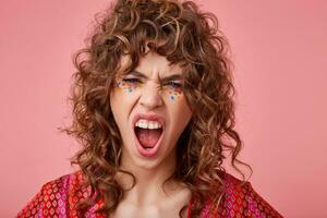 Angry young curly brunette female with festive makeup screaming violently with wide mouth opened and frowning her face, posing over pink background in colored patterned top photo