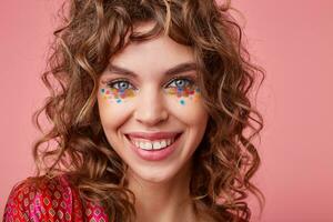 Charming young blue-eyed brunette female with multicolored dots on her face looking at camera happily and demonstrating her perfect white teeth, posing over pink background photo