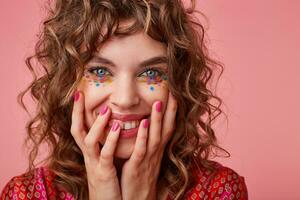 Portrait of cheerful young curly woman with festive makeup holding her face with hands and looking at camera with wide sincere smile, standing over pink background photo