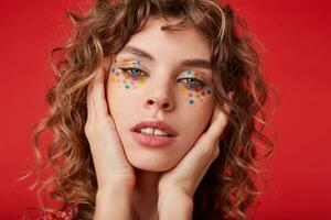 Attractive young blue-eyed woman with curly brown hair wearing festive makeup while posing over red background, holding her head with raised palms and looking at camera with calm face photo