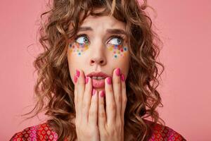 Frightened curly blue-eyed brunette female with multicolored dots on her face keeping hands on her face and looking scaredly aside, isolated over pink background photo