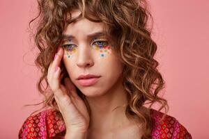 retrato de infeliz joven Rizado mujer con festivo maquillaje conmovedor suavemente su cara y mirando lejos con vacío ojos, posando terminado rosado antecedentes en de colores estampado parte superior foto