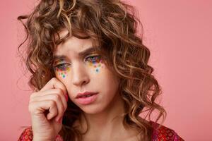 Studio photo of upset brunette curly lady with festive makeup posing over pink background, wiping away the tears and looking down sadly, being in bad spirit