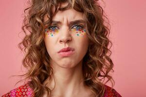 Unpleased young blue-eyed brunette woman with multicolored dots on her face frowning eyebrows and looking to camera with pout, isolated over pink background photo