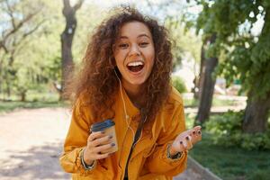 Happy broadly smiling curly darck skined girl, wearing a yellow jacket, holding a cup of coffee, enjoying the weather in the park and laughing with funny jokes, looking at the camera. photo