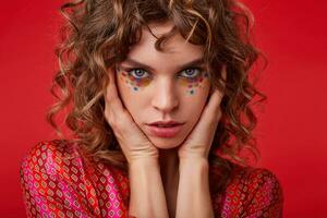 Close-up of curly blue-eyed lady with festive hairstyle and multicolored dots on her face holding neck with hands while standing over red background in colored patterened top photo