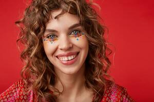 Indoor photo of charming blue-eyed young woman with romantic hairstyle wearing motley patterned top, looking at camera cheerfully and smiling widely, isolated over red background