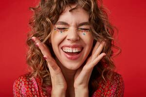 Pleasant looking curly lady with multicolored dots on her face laughing happily and showing her perfect white teeth, keeping eyes closed and raising palms to her face, standing over red background photo