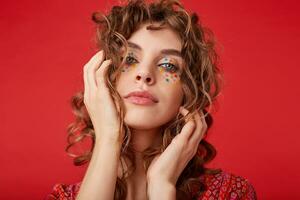 Indoor shot of lovely young lady with romantic hairstyle wearing motley patterned top, looking at camera with folded lips and keeping palms on her head, posing over red background photo