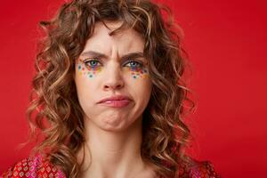 Displeased curly blue-eyed female with multicolored dots on her face twisting her mouth with frowning eyebrows, standing over red background with dissatisfied grimace photo