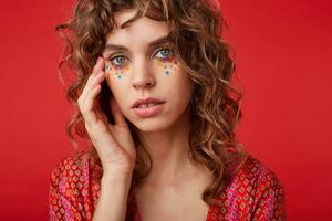 Close-up of young curly woman with multicolored dots on her face touching her face with palm gently, looking at camera with calm face, standing over red background photo