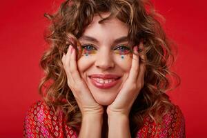 Close-up of happy pretty female with romantic hairstyle and festive makeup looking at camera with broad charming smile, holding head with raised hands while standing over red background photo