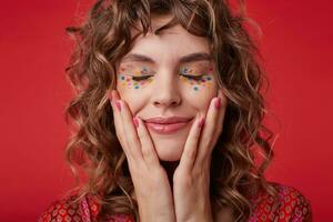 Pleasant looking pretty woman with curly hair smiling sincerely and keeping her eyes closed, holding her face with raised hands, wearing festive makeup while posing over red background photo