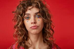 Portrait of beautiful blue-eyed young woman with romantic hairstyle and multicolored dots on her face looking to camera and pursing lips, isolated over red background photo