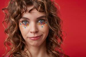 Close-up of charming blue-eyed young lady with curly hair and festive make up posing over red background, looking at camera with folded lips and calm face photo