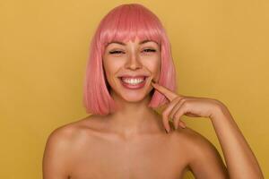 alegre joven atractivo dama con corto rosado Corte de pelo demostración su blanco Perfecto dientes mientras sonriente extensamente a cámara, conmovedor su cara con elevado índice dedo mientras posando terminado mostaza antecedentes foto